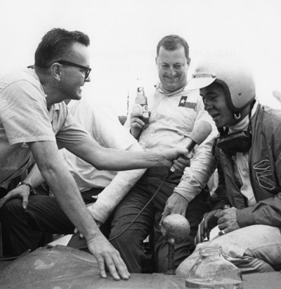 In typical fashion, Economaki interviews Roger Penske, winner of the Nassau Trophy race on Dec. 6, 1964, as teammate Hap Sharp looks on. [Image from IMRRC’s National Speed Sport News Collection]